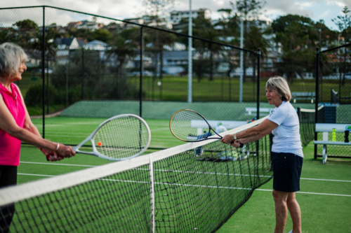 Two older women engage in a friendly tennis match at an outdoor court during the day - Australian Stock Image