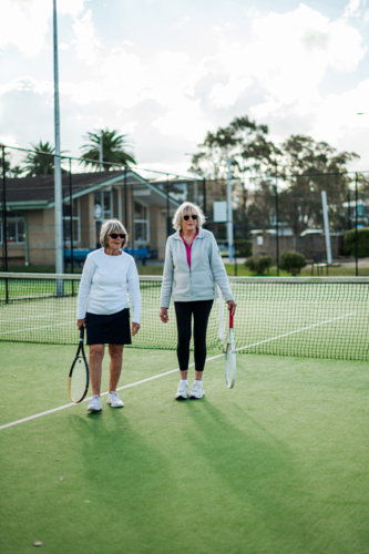 Two older women holding tennis racquets stand on a bright outdoor court in Australia. - Australian Stock Image