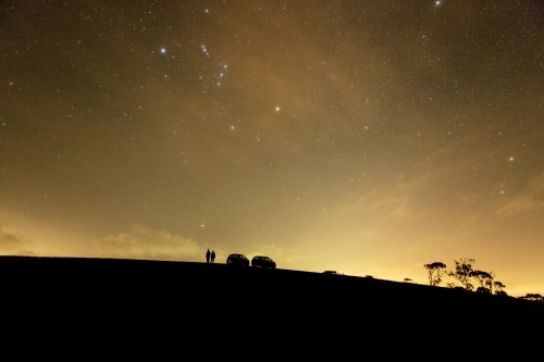 two people and two cars silhouetted on hill under stars