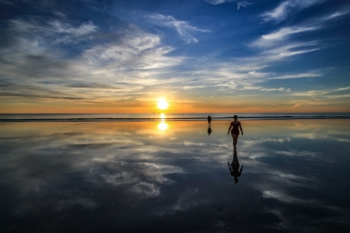 Two people walking along beach with sky reflected in water - Australian Stock Image