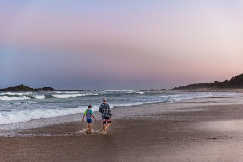 Two people walking in water on sand at beach at sunset from behind - Australian Stock Image