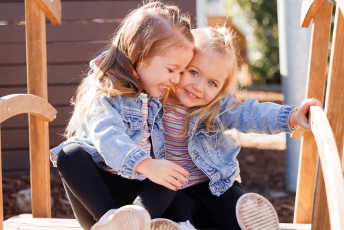 Two preschooler sisters sitting together on wooden bridge in kindergarten yard - Australian Stock Image