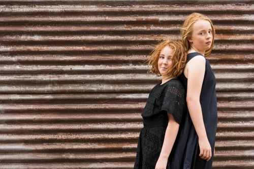 two red headed girls in black dresses back to back against rusty brown corrugated iron shed
