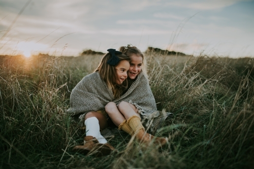 Two sisters sitting in paddock wrapped in blanket at sunset - Australian Stock Image