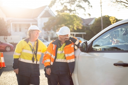 Two smiling women road workers with white helmet leaning on a white car - Australian Stock Image