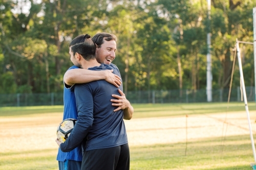 Two smiling young men on the soccer field with a soccer ball hugging each other - Australian Stock Image