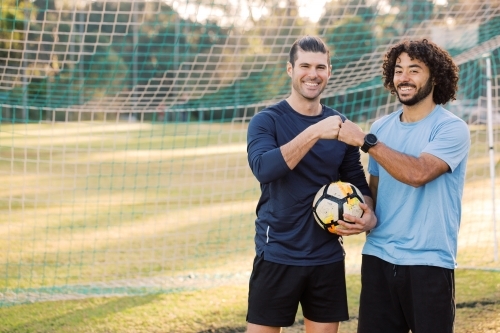 Two smiling young men standing on the field bumping their fists near the soccer net