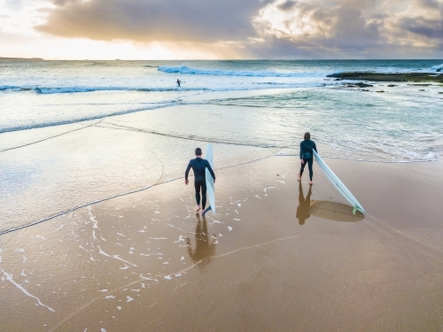 Two surfers walking on a wet beach into the ocean at sunrise