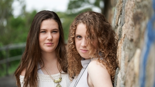 Two teen girls leaning against stone wall outdoors - Australian Stock Image
