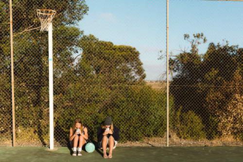 Two teenage girls on smart phones at regional outdoor netball court with ball - Australian Stock Image