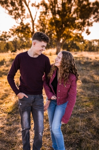 Two teens looking at each other and laughing standing in paddock at golden hour - Australian Stock Image