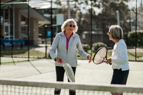 Two women in their seventies enjoy a friendly tennis match at an outdoor court, smiling joyfully - Australian Stock Image