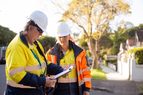 Two women road workers with white hat wearing yellow and orange jackets looking down at their notes
