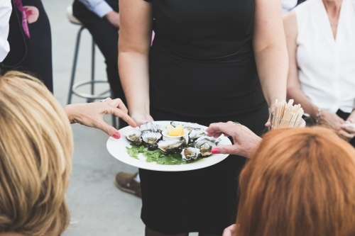 Two women's hands reaching for oysters from a platter being served by a waitress