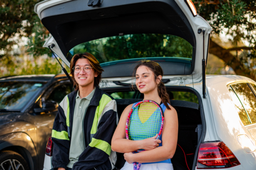 Two young adults relax by a car boot, ready to play tennis in a sunny park setting - Australian Stock Image