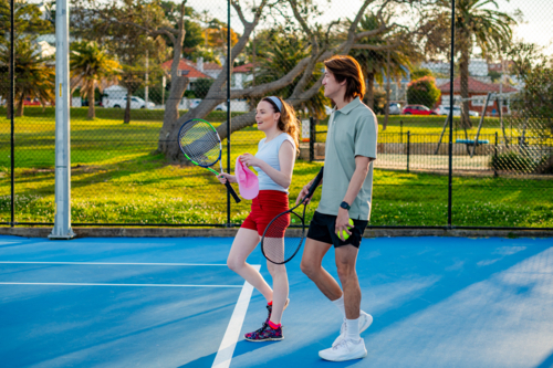 Two young adults walk together on a blue tennis court, ready for a friendly game - Australian Stock Image
