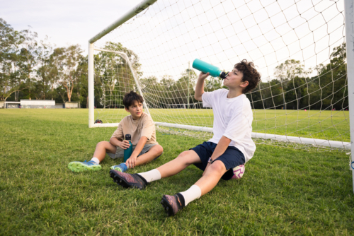 Two young boys drink from their water bottles while sitting on grassy field in front of soccer goal - Australian Stock Image