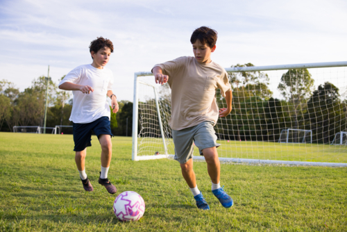 Two young boys playing soccer on grassy field near soccer goal - Australian Stock Image