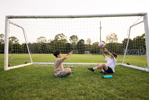 Two young boys playing with soccer ball while seated on grass field near goalpost - Australian Stock Image