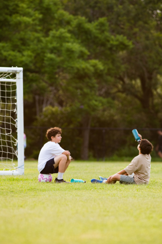 Two young boys sitting on grassy field in front of soccer goal one drinking from his drink bottle - Australian Stock Image