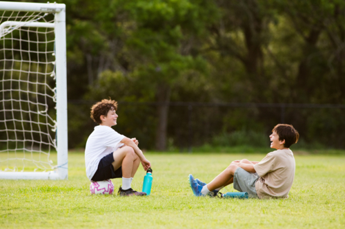 Two young boys sitting on the grassy field in front of soccer goal with their water bottles - Australian Stock Image