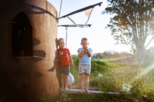 Two young boys stand next to a homemade water tank cubby house as afternoon sunlight shines in.