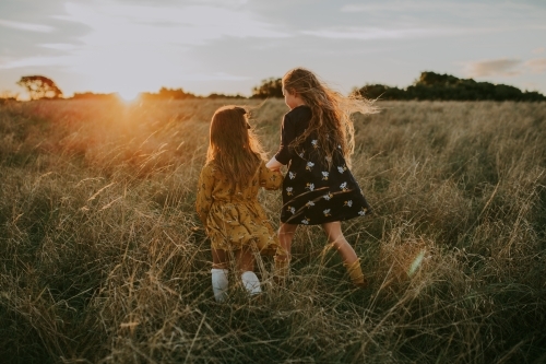 Two young fashionable sisters walking through long grass in a paddock at sunset