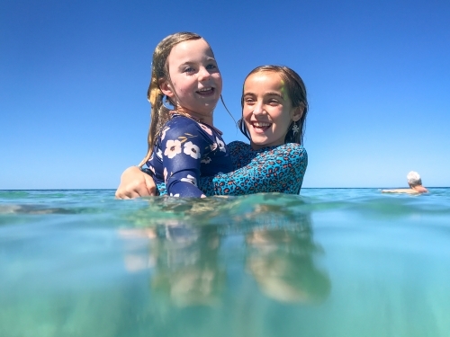 Two young girls smiling & embracing in ocean on clear sunny day in rashvests - Australian Stock Image