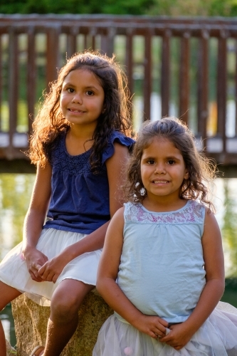 Two young girls with curly hair sitting on a huge rock outdoors looking at camera together - Australian Stock Image