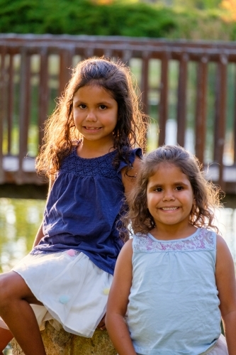 Two young girls with curly hair sitting on a huge rock outdoors looking at camera together - Australian Stock Image