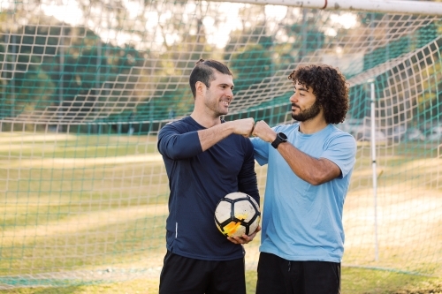 Two young men standing on the field looking at each other, bumping their fists with a soccer ball - Australian Stock Image