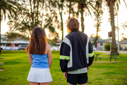 Two young people stand together, facing the sunset in a green park full of palm trees - Australian Stock Image