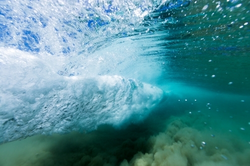 Under water view of an ocean wave breaking in a vortex on sand in crystal clear, blue water.