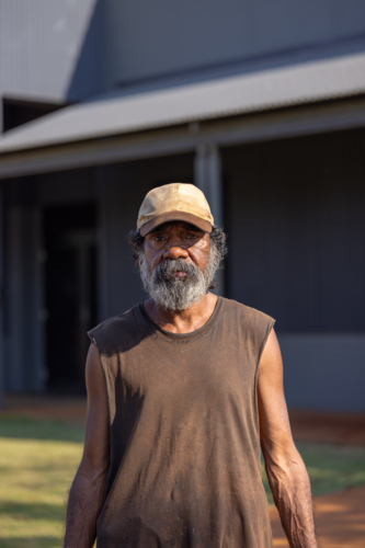upper body of Aboriginal man with greying beard standing outside wearing a cap and singlet - Australian Stock Image
