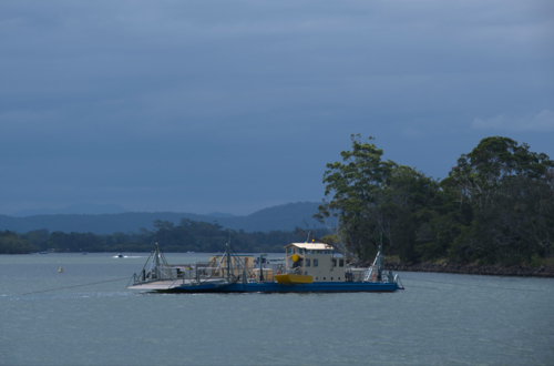 Vehicle ferry crossing river in sun, clouds and trees in background - Australian Stock Image