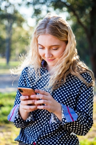 Vertical portrait of a happy young woman texting on her mobile phone outside - Australian Stock Image