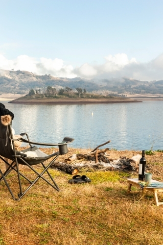 An extinguished campfire with foldable chair pair of slippers and wine beside the lake - Australian Stock Image