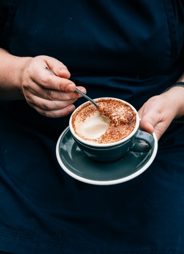 Vertical shot of a hand holding a saucer and a cup of coffee while scraping off the froth in it. - Australian Stock Image
