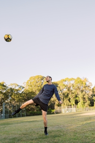 vertical shot of a man kicking a ball up in midair with one leg raised with trees in the background - Australian Stock Image