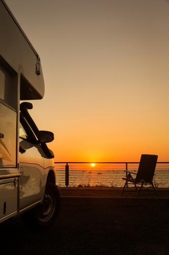 Vertical shot of a white camping van during sunset in front of the sea with foldable chair - Australian Stock Image