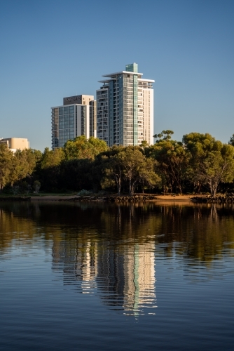 vertical shot of buildings with trees and reflections appearing in a lake under clear blue skies