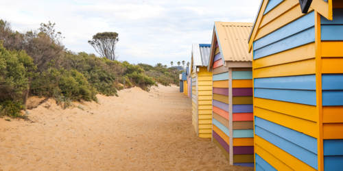 Vibrant beach huts on sandy path with greenery and cloudy sky in background - Australian Stock Image