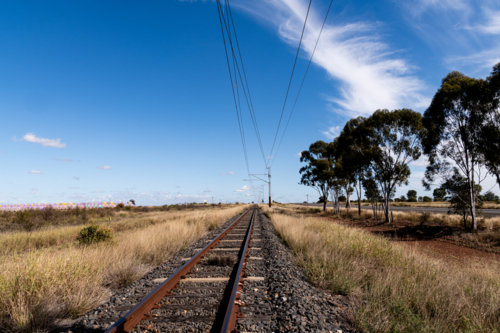 view along railway tracks lined with dry grass and trees and cotton bales in distance - Australian Stock Image
