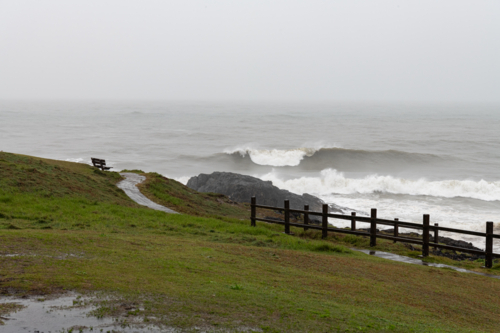 View from a headland of waves breaking in a grey ocean in rainy wild weather - Australian Stock Image