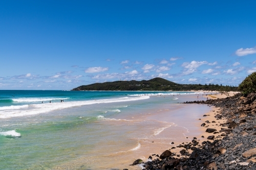 View of Cape Byron and lighthouse from the beach at Byron Bay with surf , rocks and sandy foreshore - Australian Stock Image