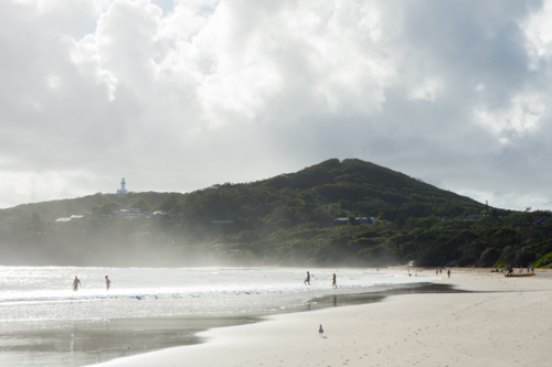 View of Cape Byron and lighthouse from the beach with swimmers and sandy foreshore - Australian Stock Image