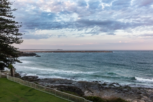 View of ocean beach with rocks and tree and cloudy sky - Australian Stock Image