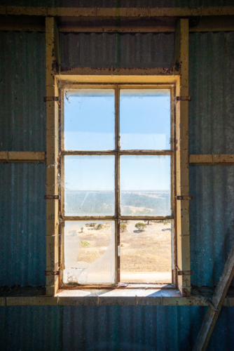 View through a cracked window in an old corrugated iron building overlooking a dry rural landscape. - Australian Stock Image