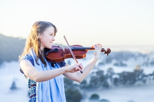 Violin player standing out on cliff top playing music on misty morning - Australian Stock Image