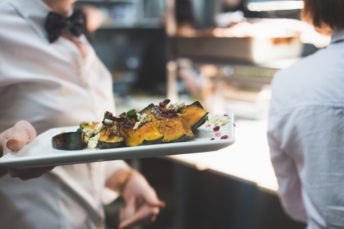 Waiter wearing a bow tie and carrying a plate of roasted pumpkin in a restaurant kitchen